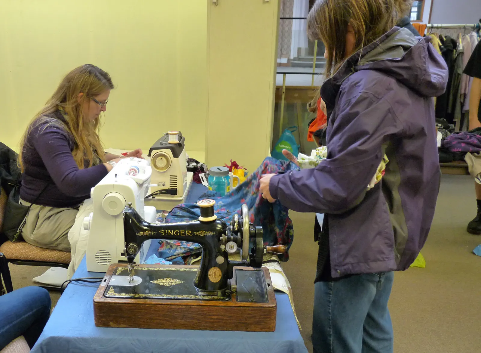 A volunteer at a sewing machine mending a garment while another woman holds up a blue floral fabric beside a black Singer hand-crank machine